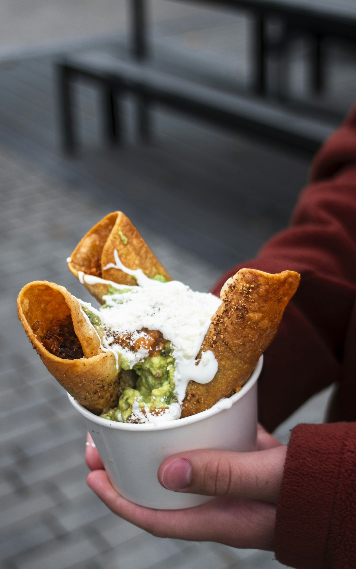 A cup of crispy rolled taquitos held in one hand, topped with guacamole, crema and grated cheese